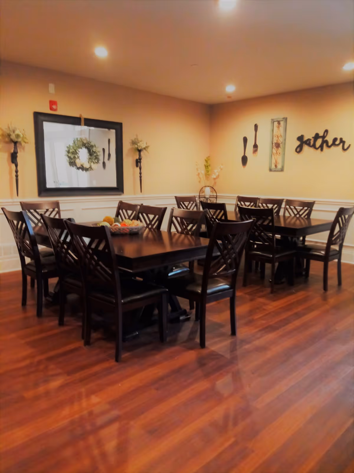 Dining room with several dark wood tables and chairs on a polished hardwood floor, a wall mirror and decorative wall art including a 'gather' sign.