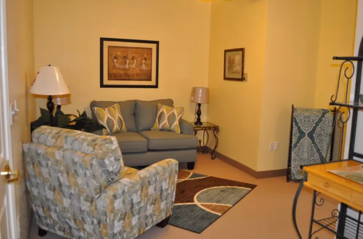 A cozy living room with a gray loveseat adorned with patterned pillows, a patterned armchair, two table lamps on side tables, framed artwork on yellow walls, a decorative rug on the floor, and a small wooden table with a metal frame in the corner.