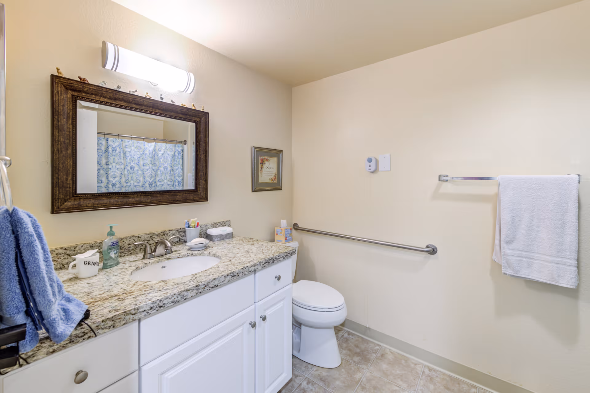 A clean bathroom with a granite countertop, white cabinets, a sink with a faucet, a large mirror with a dark wooden frame, a toilet with a grab bar beside it, a towel rack with a white towel, and a blue patterned shower curtain reflected in the mirror.