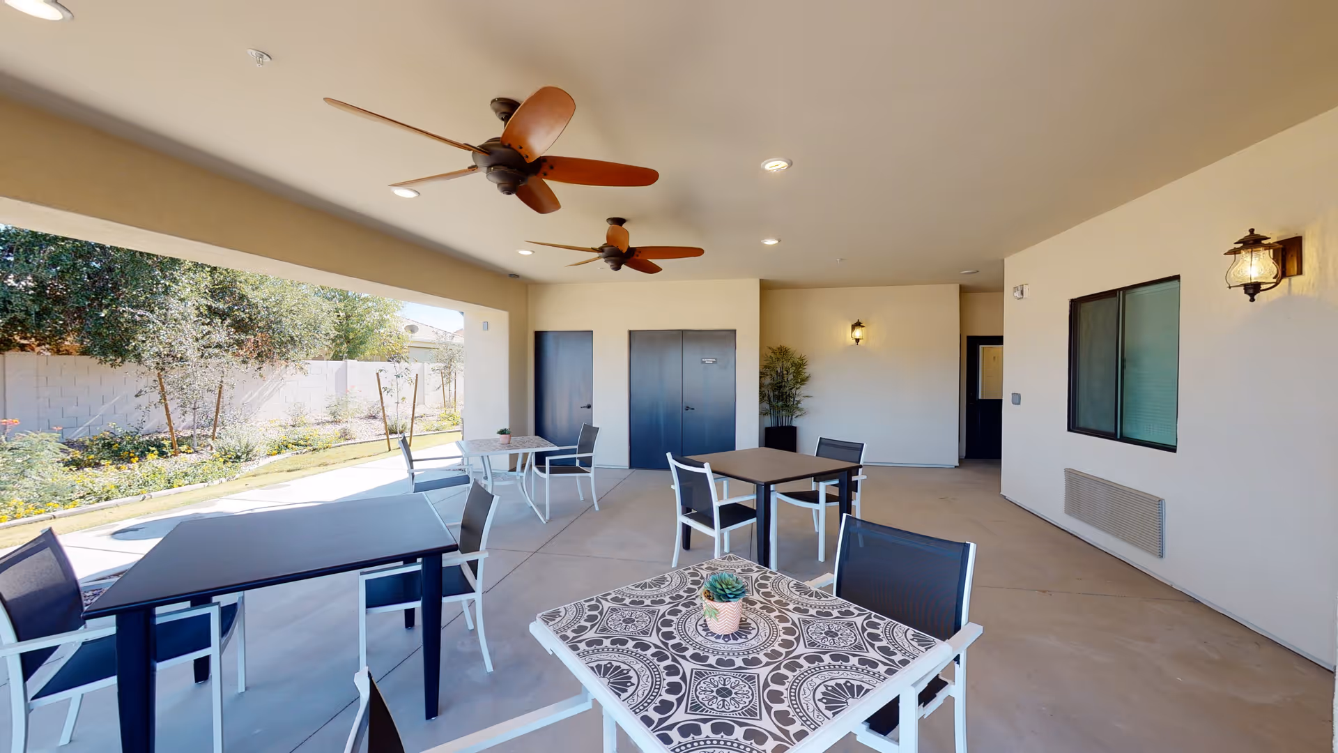 Covered outdoor patio with several dining tables and chairs, ceiling fans, and a view of a garden.