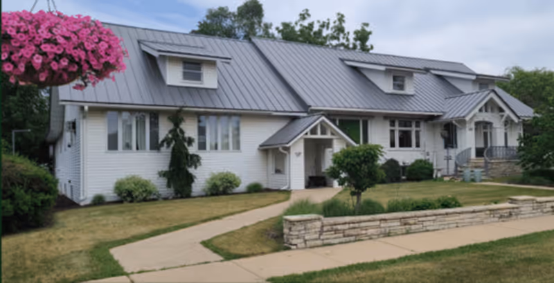 Exterior view of a single-story white building with a metal roof, surrounded by a lawn and a stone retaining wall. There is a hanging basket with pink flowers on the left side and a concrete walkway leading to the entrance.
