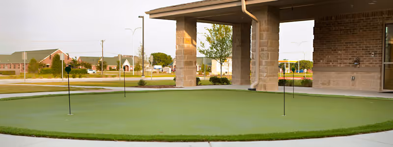 Outdoor putting green area with multiple golf holes and flags, adjacent to a covered patio structure with stone pillars. In the background, there are buildings, trees, and a road under a clear sky.