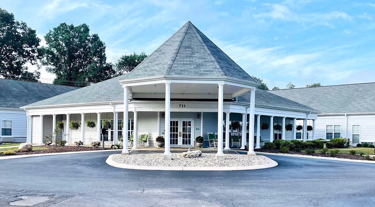 Front entrance of a single-story senior living building with a covered circular drive, white columns, rocking chairs, and hanging plants.