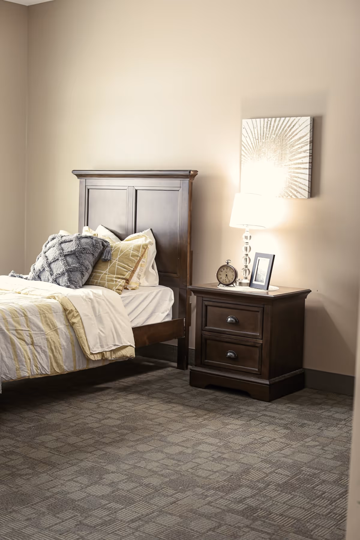 A bedroom with a wooden bed frame and a neatly made bed with white and yellow bedding and multiple pillows. Next to the bed is a wooden nightstand with two drawers, a table lamp, an alarm clock, and a framed photo. A decorative wall art piece hangs above the nightstand. The room has beige walls and patterned carpet flooring.
