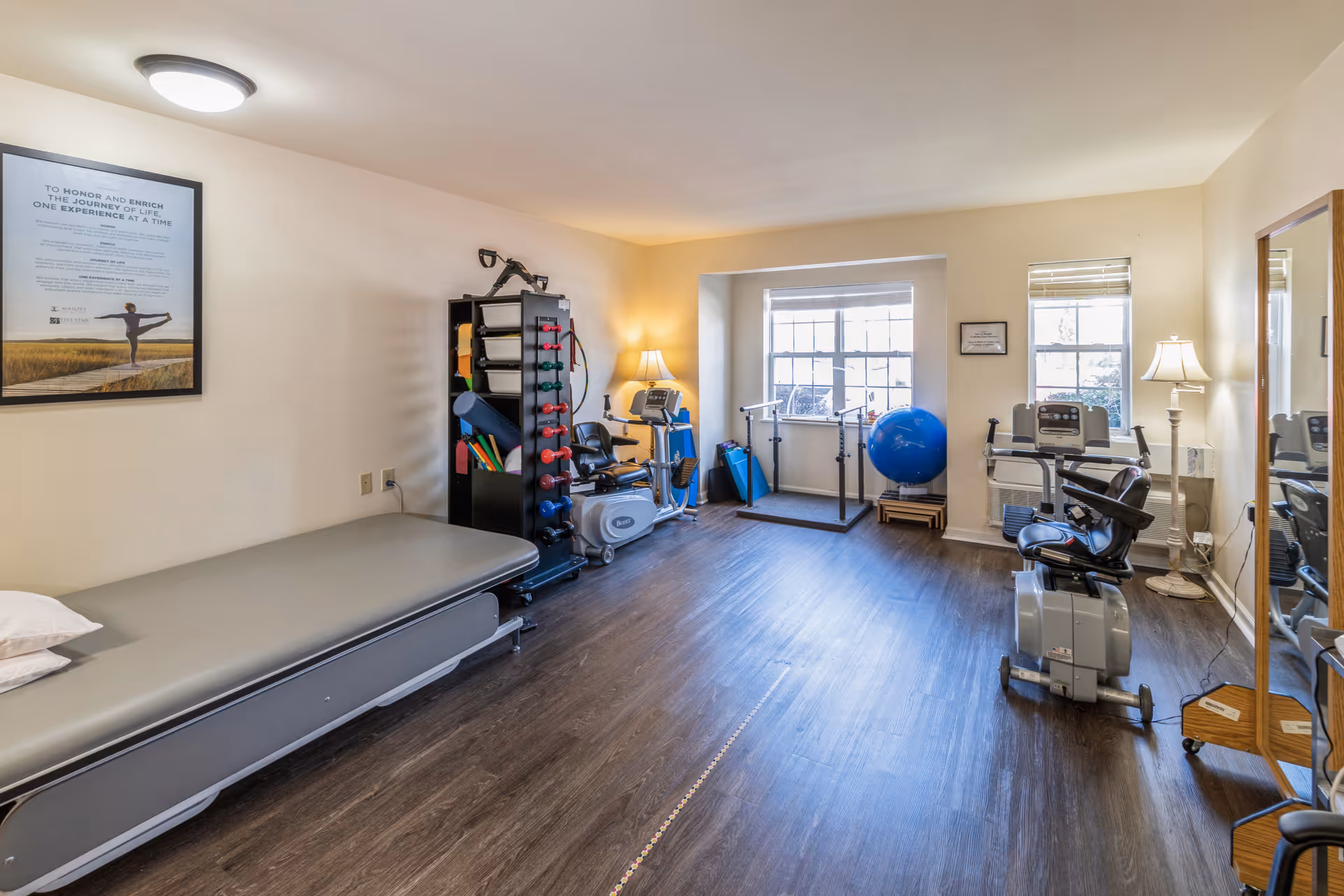 A well-lit exercise room with wooden flooring featuring various fitness equipment including a recumbent exercise bike, a rack with colorful dumbbells and exercise mats, a large blue exercise ball, and a ballet barre near the windows. There is a padded bench with pillows on the left side and a full-length mirror on the right side. The room has beige walls with a framed motivational poster and two standing lamps providing additional light.