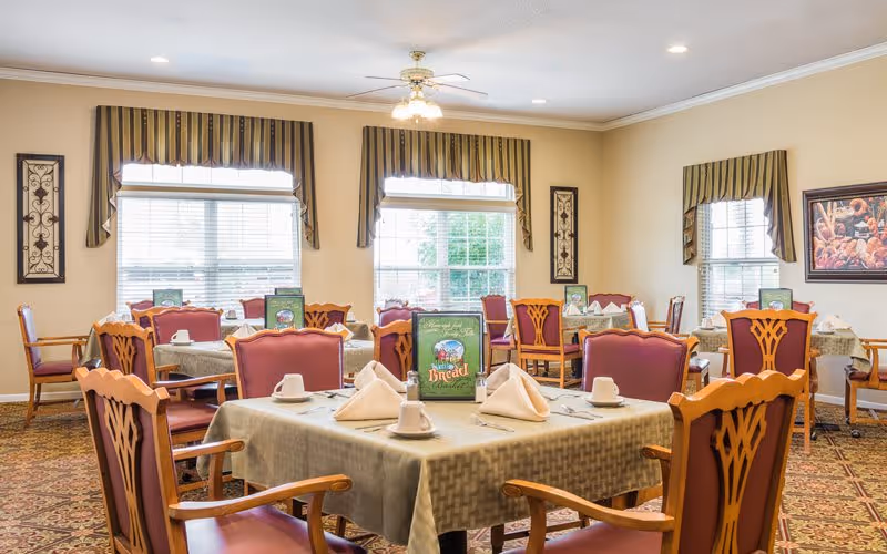 A bright dining room with multiple tables covered in beige tablecloths, each set with white napkins, cups, and silverware. The chairs have wooden frames with red cushioned seats and backs. The room has large windows with striped valances, framed artwork on the walls, and a patterned carpet.