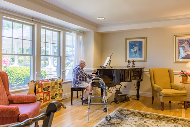 An elderly man playing a black grand piano in a well-lit living room with large windows. The room features two upholstered armchairs, a colorful quilt draped over a wooden rack, framed artwork on the walls, and a patterned area rug on the wooden floor. A walker is positioned near the man.