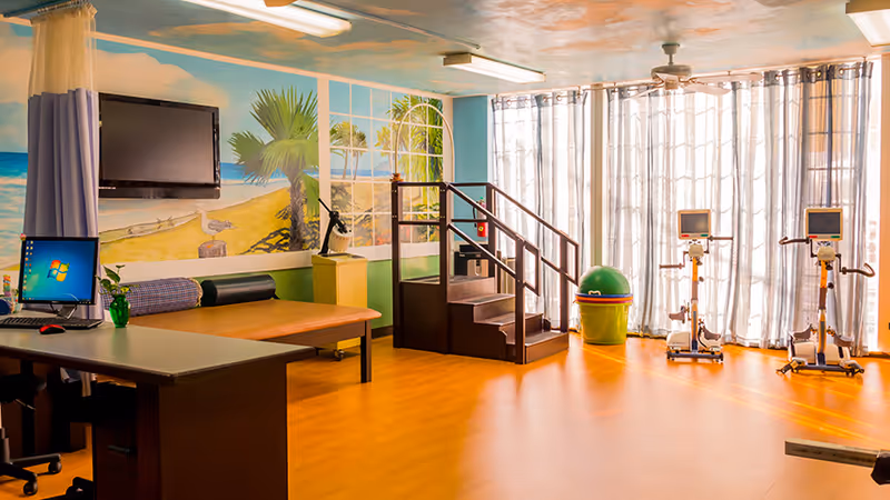 Bright indoor senior fitness/rehab room with exercise bikes, a stair-step platform, a mural beach wall, and a desk with a computer.