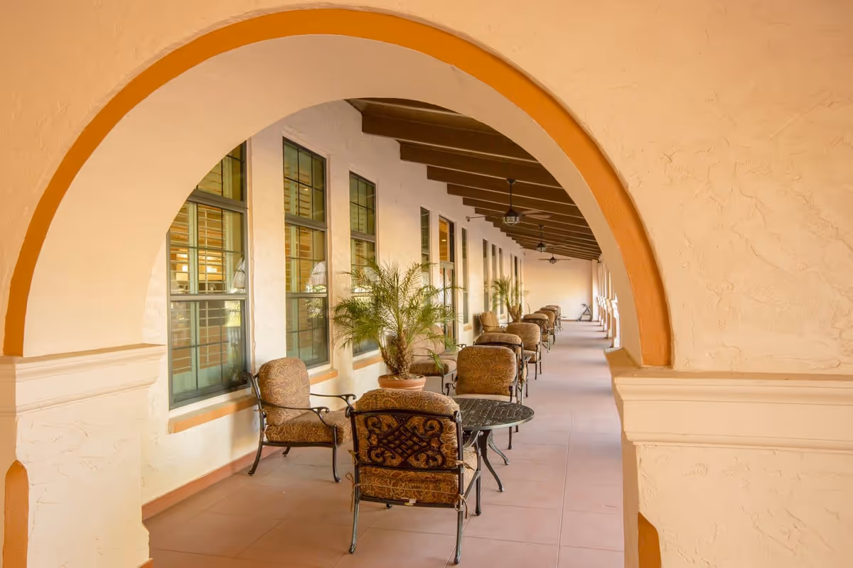 Covered outdoor corridor with arched openings, lined with cushioned chairs and small round tables, potted plants, and ceiling fans under a wooden beam ceiling.