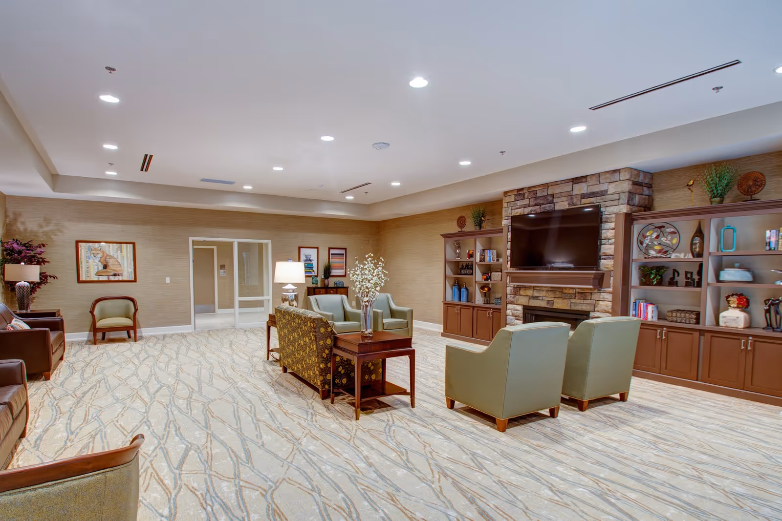 A spacious senior living facility common area with multiple armchairs and sofas arranged around a wooden coffee table. The room features a stone fireplace with a mounted flat-screen TV above it, flanked by built-in wooden shelves filled with decorative items and books. The walls are beige with framed artwork, and the floor is carpeted with a light pattern. The ceiling has recessed lighting, and there is a glass door leading to another area.