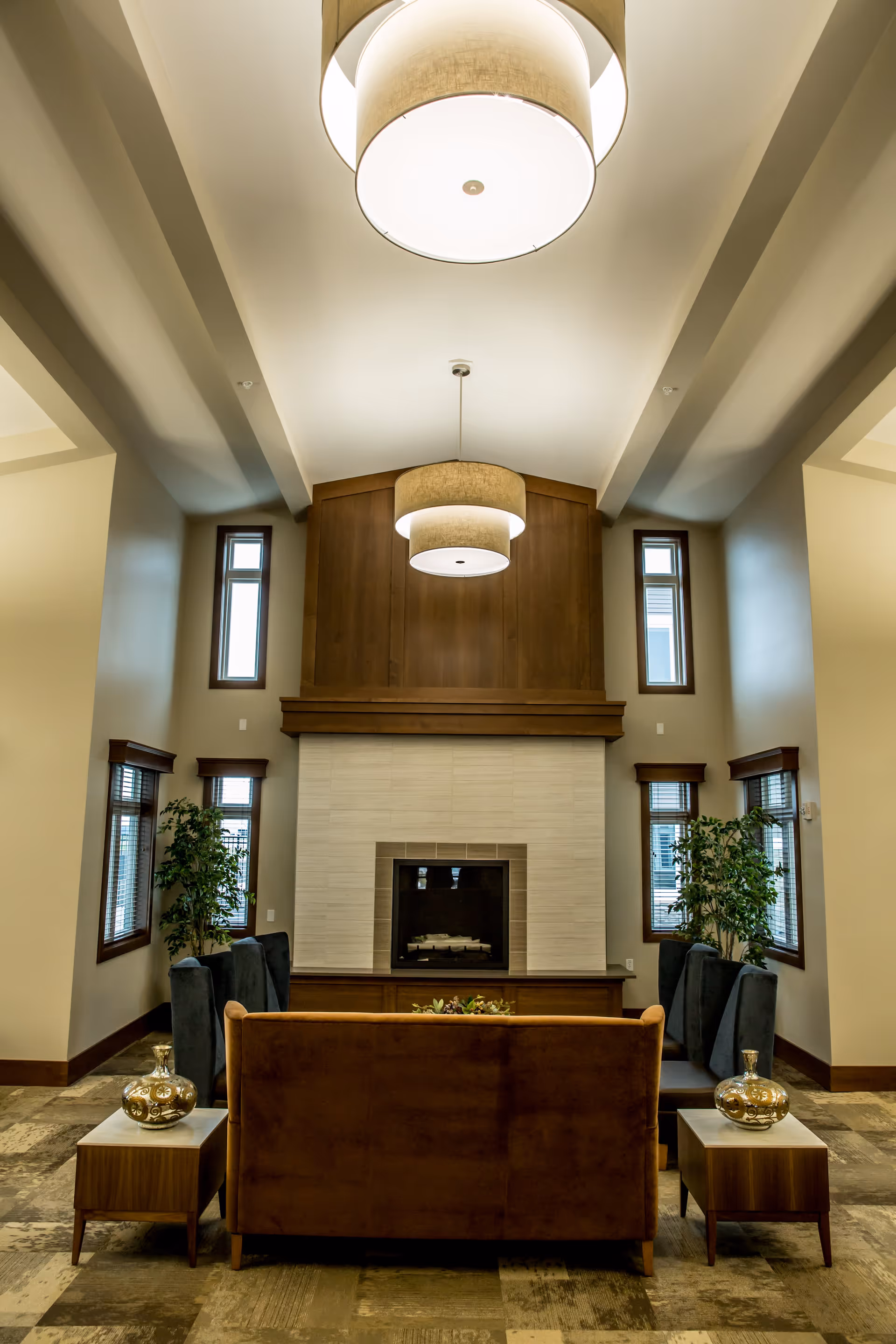 A cozy living room area with a brown sofa facing a modern fireplace with a wooden mantel and tiled surround. The room has tall ceilings with two large circular pendant lights hanging down. There are two side tables with decorative vases on either side of the sofa, and four dark upholstered chairs arranged around the fireplace. The walls are light-colored with several windows framed in dark wood, and two tall green plants are placed near the windows.