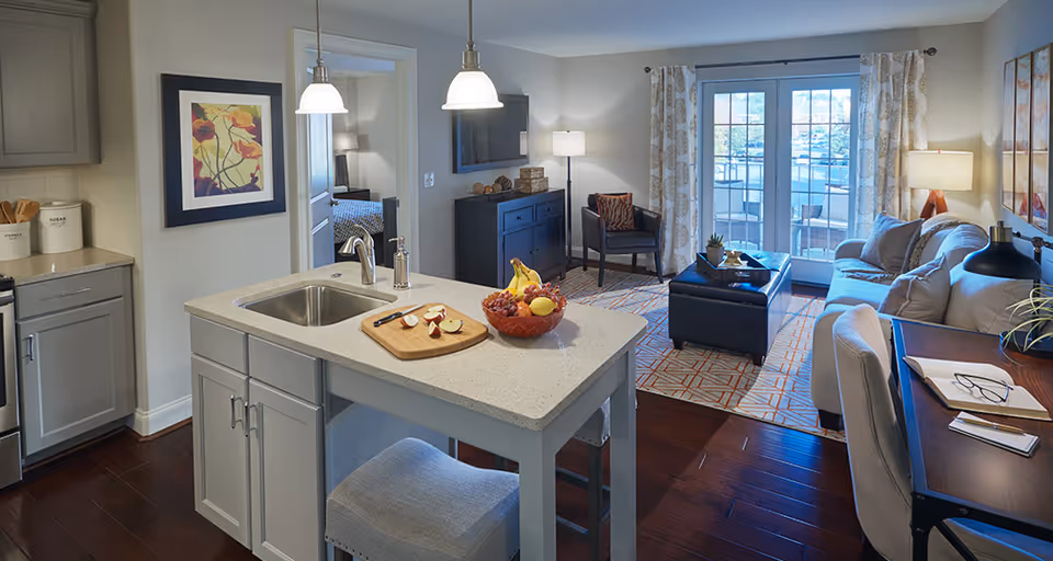 Open-plan senior living apartment with a kitchen island in the foreground and a furnished living room with sofa, coffee table, and balcony doors in the background.
