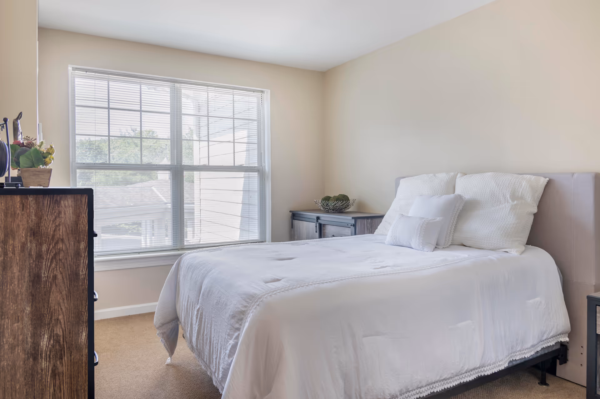 A bright bedroom with a large window letting in natural light. The bed is made with white bedding and several pillows. There is a wooden dresser on the left side and a small cabinet with a decorative bowl on the right side of the bed. The walls are painted a light beige color.
