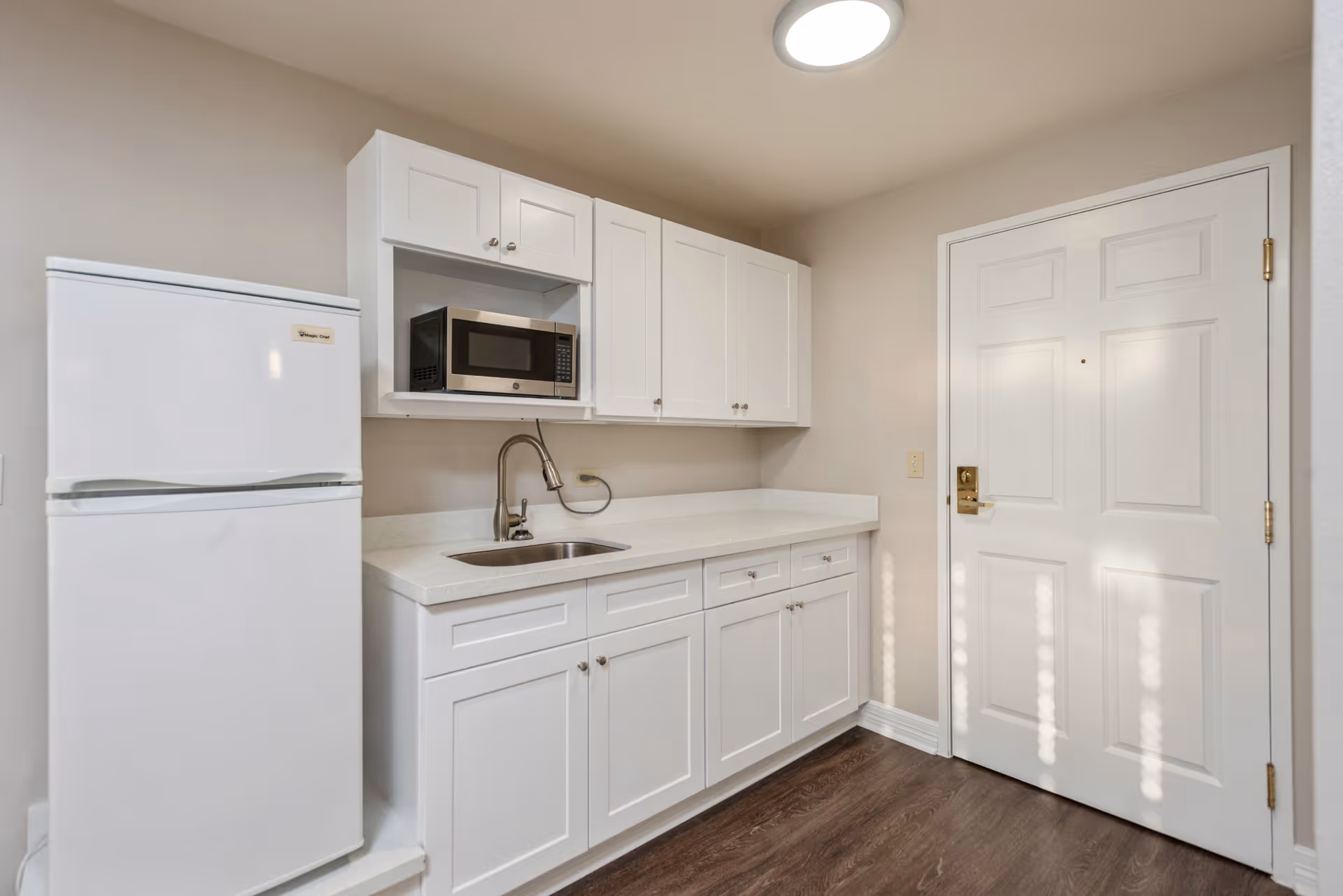 A small kitchen area with white cabinets, a white refrigerator, a microwave oven, a stainless steel sink with a faucet, and a white door on the right side. The floor is dark wood, and the walls are painted beige.