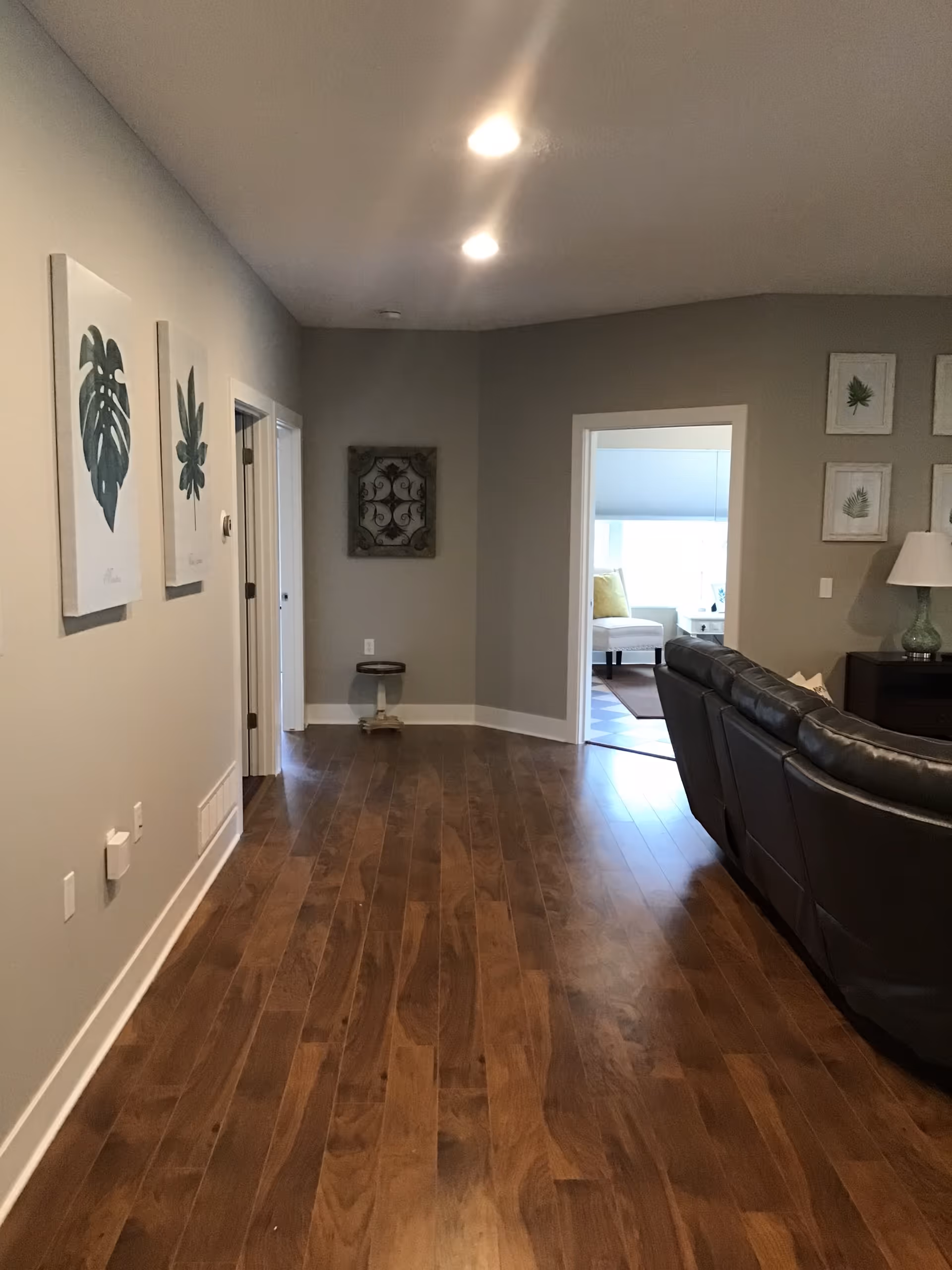 Interior view of a senior living facility hallway with wooden flooring, beige walls decorated with framed botanical prints, and a partial view of a living room area with a dark leather sectional sofa and a lamp on a side table.