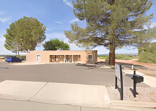 Single-story beige building with large windows set behind a paved driveway and mailboxes, surrounded by trees.