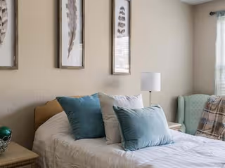 A cozy bedroom with a neatly made bed featuring white bedding and several blue and white pillows. There are three framed feather artworks hanging on the beige wall above the bed. A bedside table with a decorative item is on the left, and on the right, there is a small table with a white lamp and a light green armchair with a plaid blanket draped over it near a window with sheer curtains.