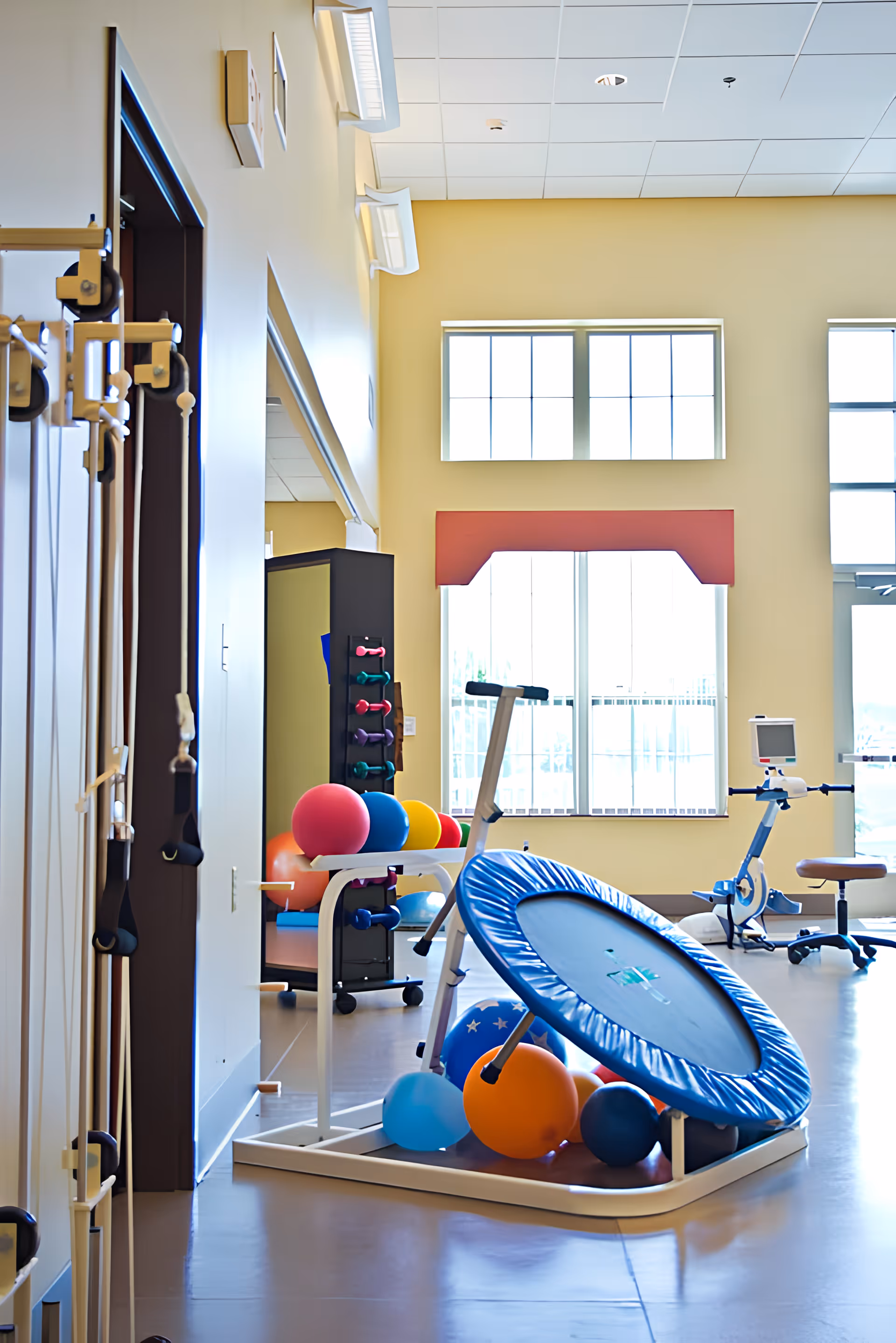 Bright physical therapy room with exercise equipment including a blue mini trampoline, colorful exercise balls, resistance bands hanging on the wall, a stationary bike, and a rack of small dumbbells. Large windows allow natural light to fill the space.