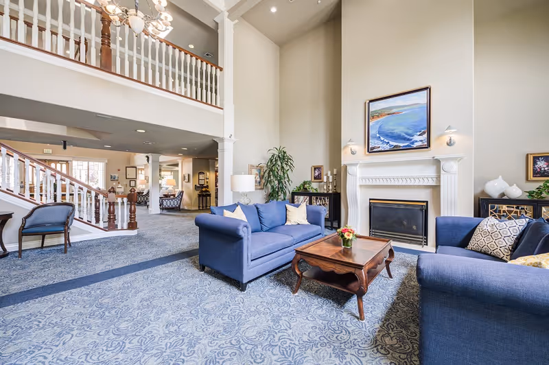 A spacious and elegant living room area in a senior living facility featuring two blue upholstered sofas with decorative pillows, a wooden coffee table with a small flower arrangement, a white fireplace with a framed coastal painting above it, and a tall plant in the corner. The room has high ceilings, patterned carpet, and a staircase with white and wooden railings leading to an upper level.