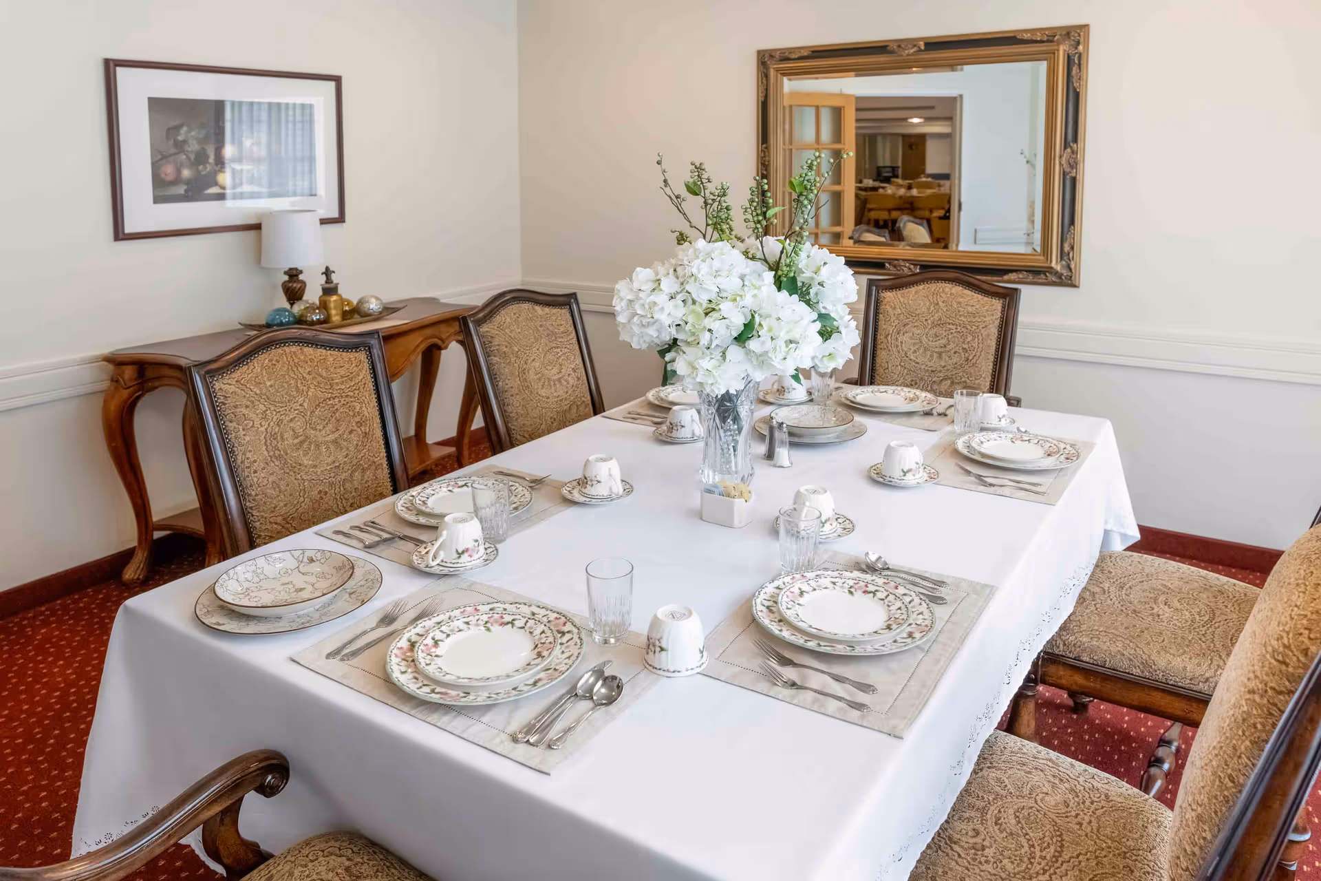 A formal dining room with a table set for six, a large floral centerpiece, ornate chairs, and a mirror on the wall.