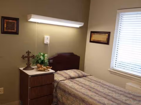 A small bedroom in a senior facility with a bed, dresser, plant, and window.