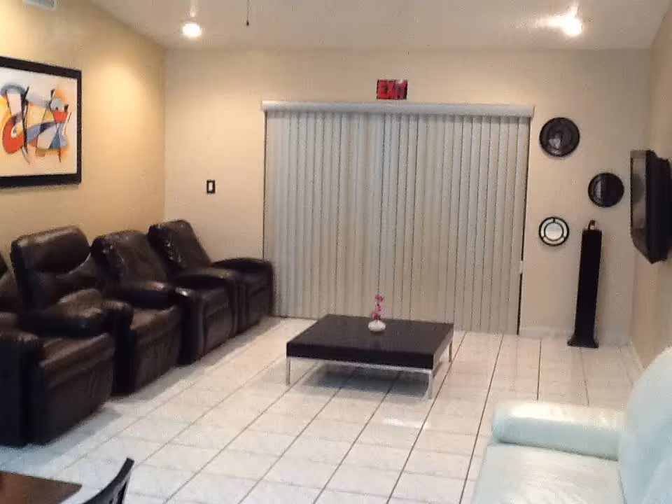 Common room with a row of dark leather recliners, a low coffee table, vertical blinds and a wall-mounted TV.