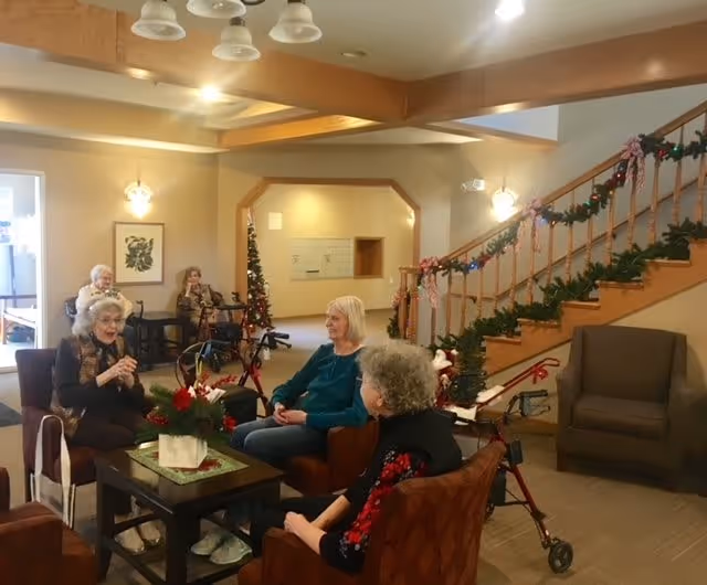 A cozy senior living facility common area decorated for the holidays with garlands and bows on the staircase railing. Four elderly women are seated in armchairs around a coffee table with a festive centerpiece, engaging in conversation. In the background, two more elderly women are seated near a window. The room has warm lighting and a welcoming atmosphere.