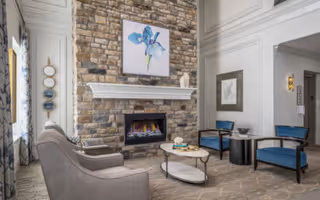 A cozy living room area featuring a stone fireplace with a colorful floral painting above it. The room includes a gray upholstered sofa, two blue cushioned chairs, a round coffee table with decorative items, and a neutral-toned carpet. The walls are light-colored with decorative molding, and there is natural light coming from a window on the left side.