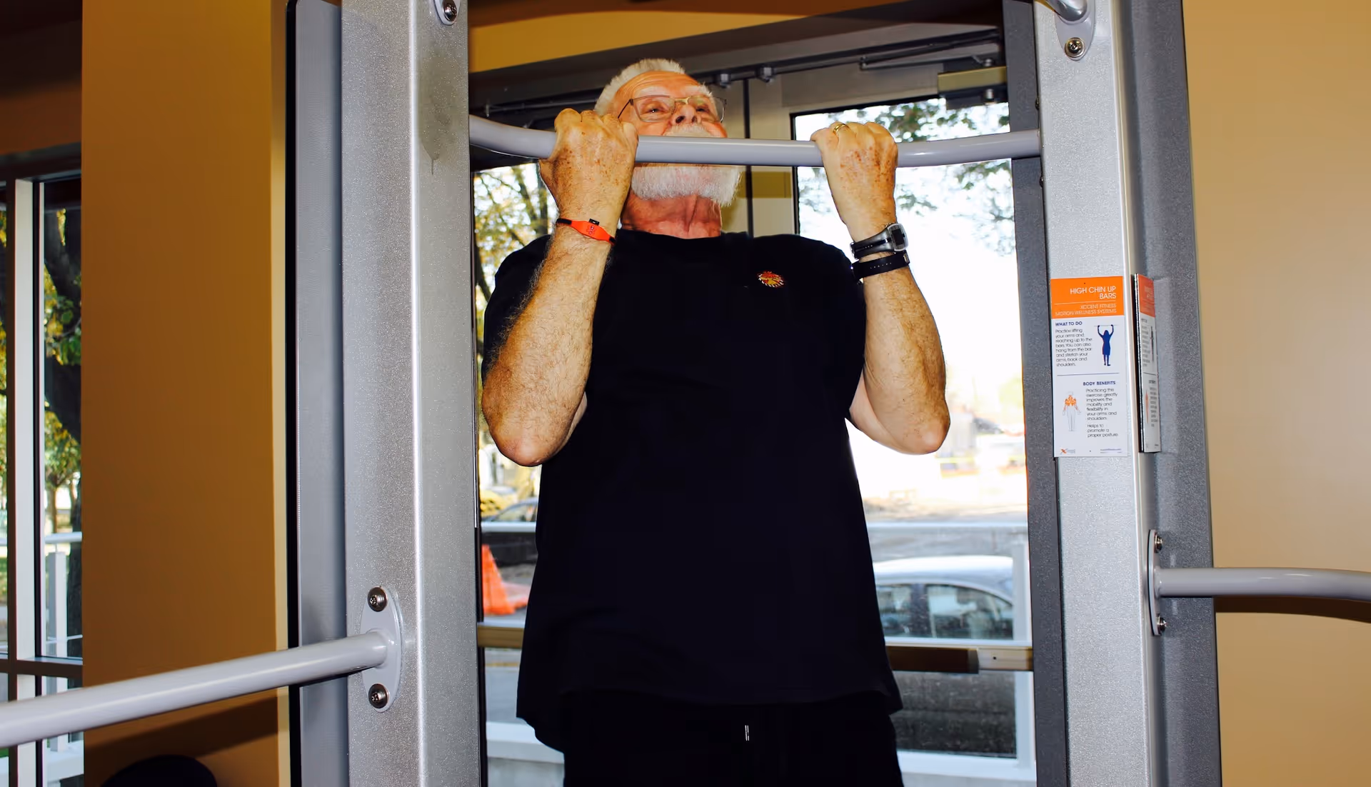 An elderly man with white hair and beard wearing glasses and a black t-shirt is doing a pull-up exercise on a high chin-up bar inside a fitness room with large windows showing trees and parked cars outside.