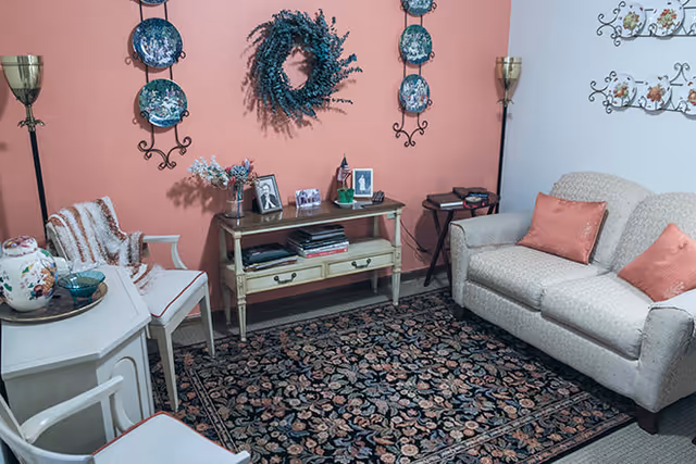 A cozy living room with a floral patterned area rug, a beige loveseat with two coral-colored pillows, two white chairs with cushions, a small wooden console table with framed photos and decorative items, a floral wreath hanging on a coral-colored wall, and two tall brass floor lamps.