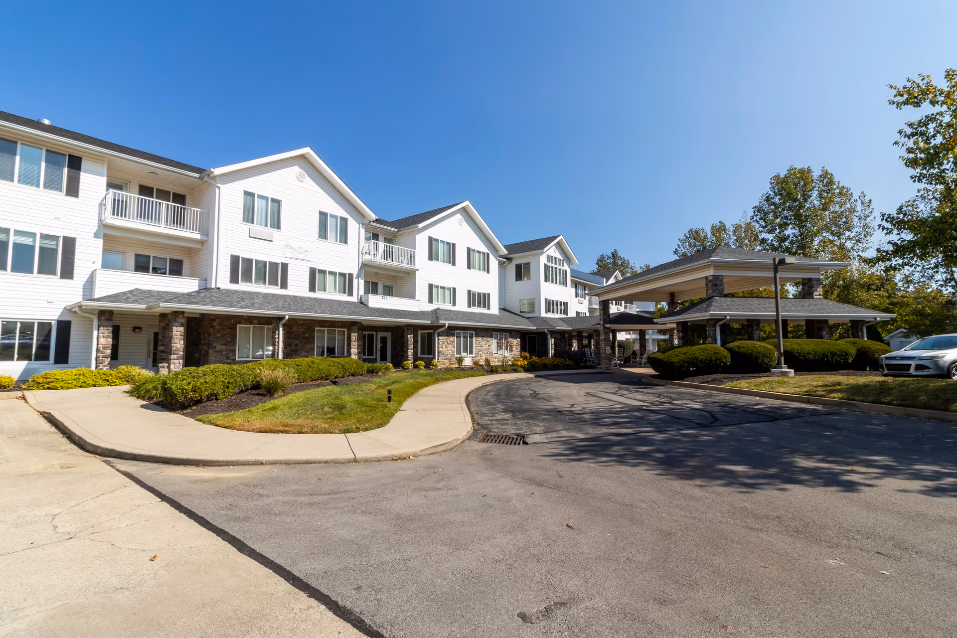 Exterior view of a multi-story senior living facility building with white siding and stone accents. The building has several windows and balconies. There is a covered driveway entrance with stone pillars and landscaped bushes around the area. A car is parked on the right side under clear blue sky.