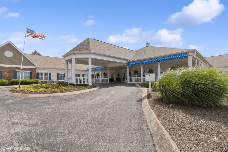 Exterior view of The Neighborhood at Hopkinsville senior living facility showing a driveway leading to a covered entrance with white pillars and blue striped awnings. There is an American flag on a flagpole to the left and landscaped bushes and plants around the driveway.