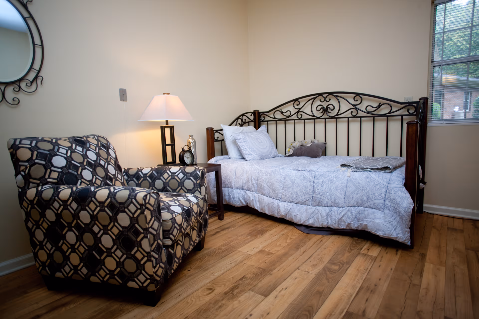 A cozy bedroom with a patterned armchair, a metal-framed daybed with pillows, a side table lamp and hardwood floors.