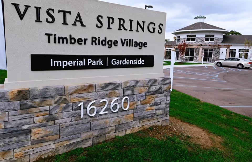 Entrance sign for Vista Springs Timber Ridge Village with the address number 16260 displayed on a stone base. In the background, there is a building with a circular section and a parking lot with a silver car parked.