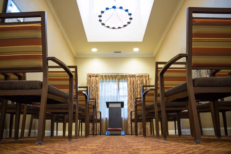 View of a small meeting or dining room with rows of wooden chairs with striped cushions facing a podium at the front. The room has a skylight with a circular chandelier, recessed ceiling lights, and a window with patterned curtains letting in natural light.