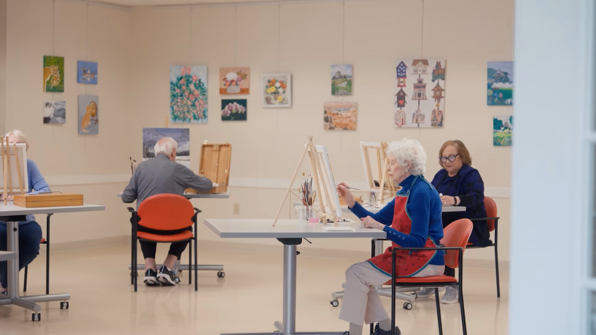 A group of elderly people seated at tables in a well-lit room, engaged in painting on canvases set on easels. The walls are decorated with various colorful paintings, and the individuals appear focused on their artwork.
