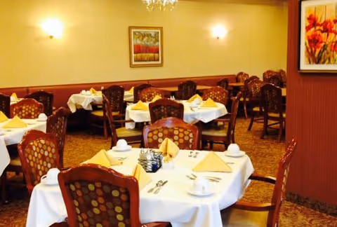 Dining room with multiple round tables covered with white tablecloths, each set with yellow folded napkins, cups, and silverware. The chairs have patterned upholstery. The walls are decorated with framed artwork and wall sconces providing warm lighting.