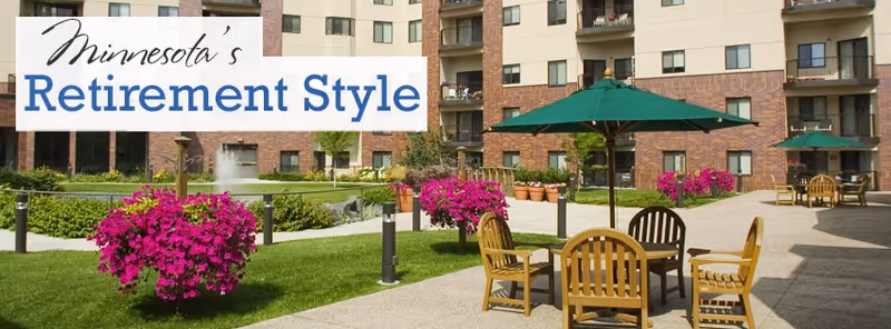 Outdoor courtyard area of a senior living facility with green grass, vibrant pink flowers in planters, wooden chairs and tables with green umbrellas, and a multi-story building in the background.