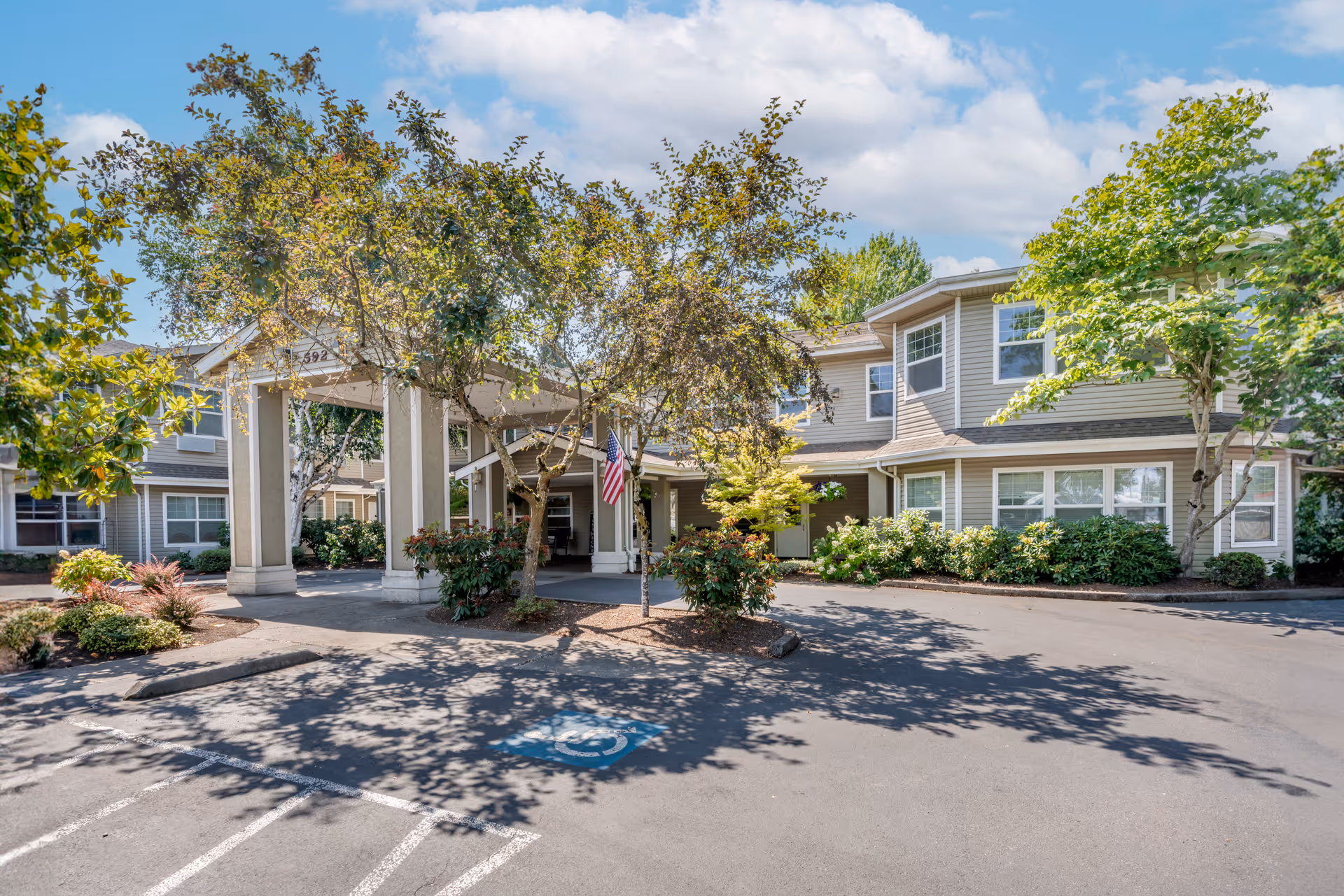 Exterior view of Brookdale River Road senior living facility showing the entrance with a covered drop-off area, surrounded by trees and landscaping under a partly cloudy sky.