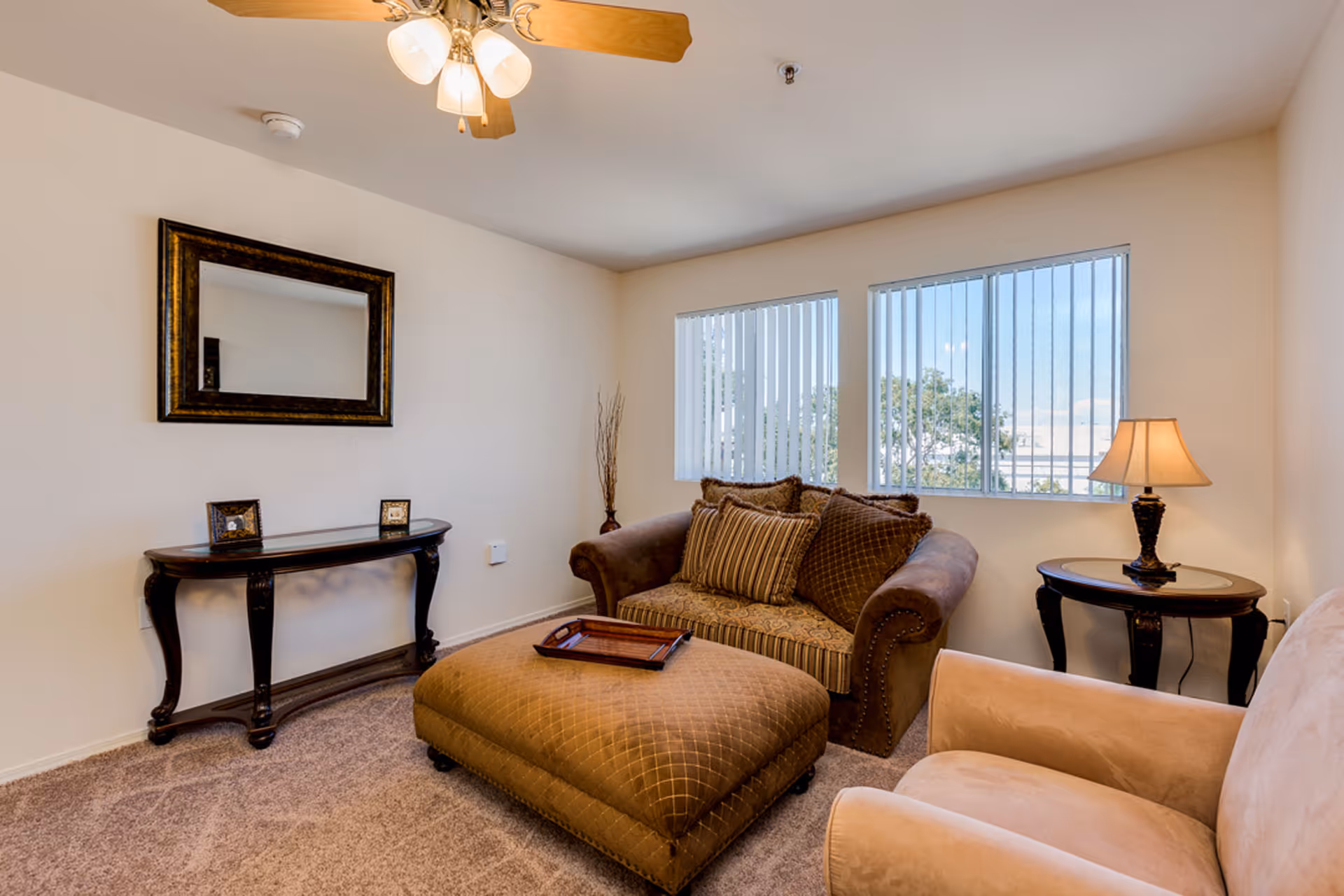 A cozy living room with a brown upholstered loveseat adorned with multiple cushions, a matching ottoman with a wooden tray on top, a beige armchair, a dark wooden side table with a lamp, a decorative console table with two small framed pictures, a large framed mirror on the wall, and two windows with vertical blinds letting in natural light.
