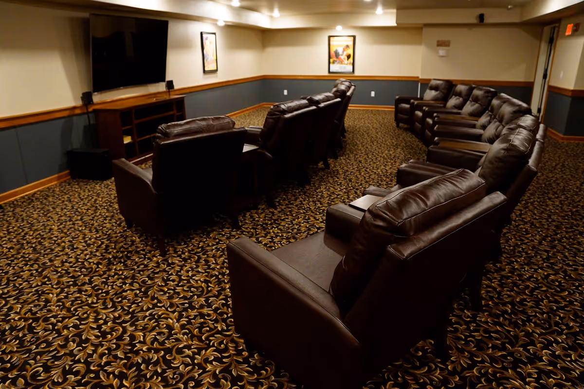 A cozy media room with two rows of brown leather recliner chairs facing a large flat-screen TV mounted on the wall. The room has patterned carpet flooring, beige walls with a blue-gray wainscoting, and framed artwork on the walls.