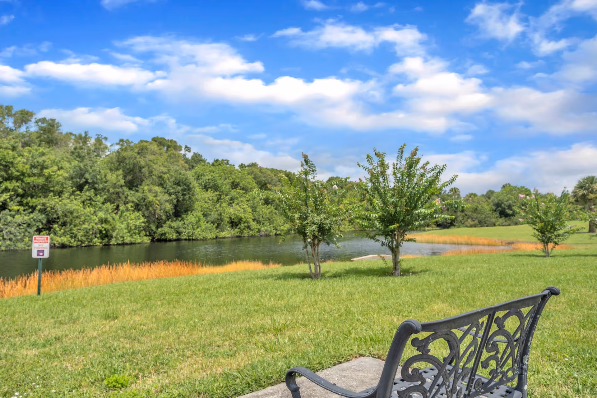 A peaceful outdoor scene featuring a small lake surrounded by lush green grass and trees under a partly cloudy blue sky. In the foreground, there is a decorative black metal bench on a concrete slab facing the water.