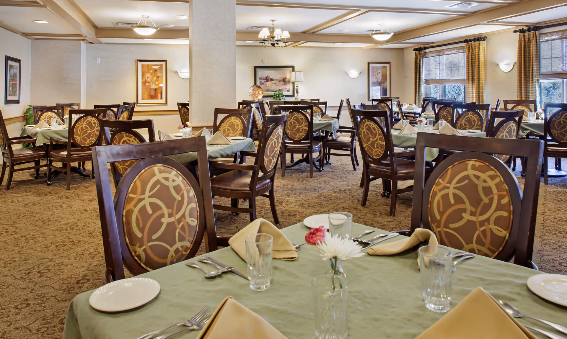 A well-lit dining room with multiple tables covered in green tablecloths, each set with beige napkins, plates, glasses, and silverware. The chairs have dark wooden frames with patterned upholstery. The room features beige walls, framed artwork, large windows with curtains, and ceiling lights.