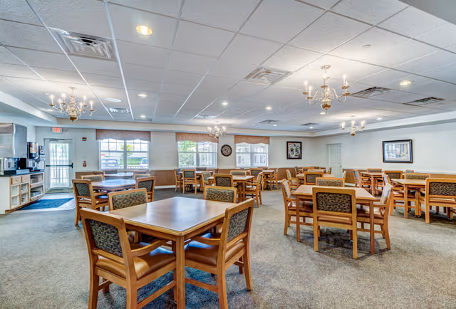 A spacious dining room with multiple wooden tables and chairs arranged neatly. The room has large windows letting in natural light, chandeliers hanging from the ceiling, and framed artwork on the walls. There is a coffee station and a door leading outside on the left side.