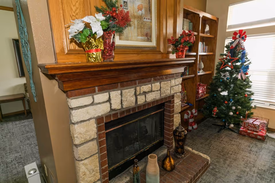 Cozy living room corner with a stone and brick fireplace decorated with holiday ornaments, including a poinsettia plant and red flowers on the mantel. To the right, there is a decorated Christmas tree with wrapped presents underneath, next to a wooden bookshelf filled with books and holiday decorations. A window with blinds allows natural light into the room.