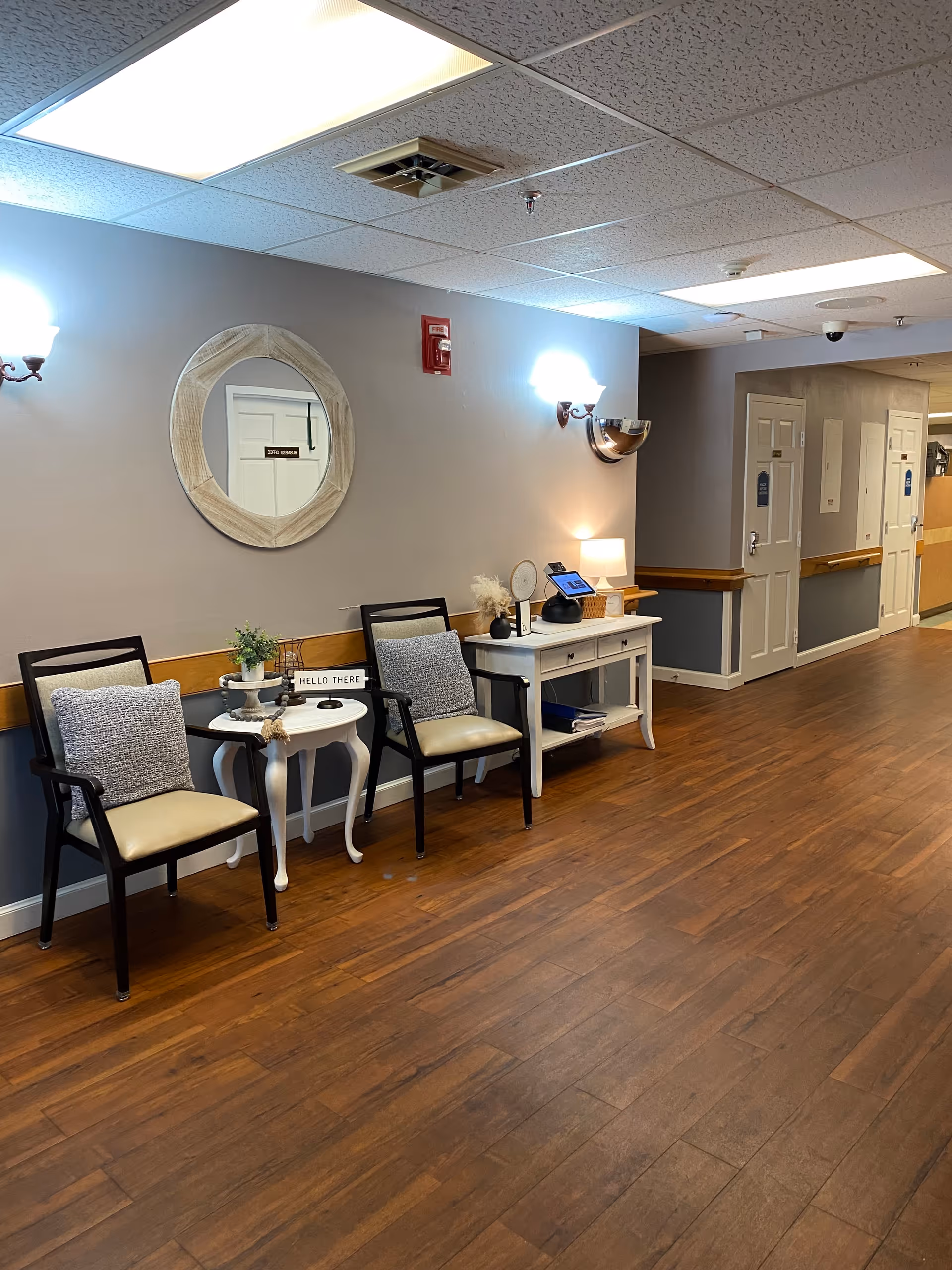 Hallway seating area with two chairs, side tables, a console table, wall mirror and sconces in a senior living facility.