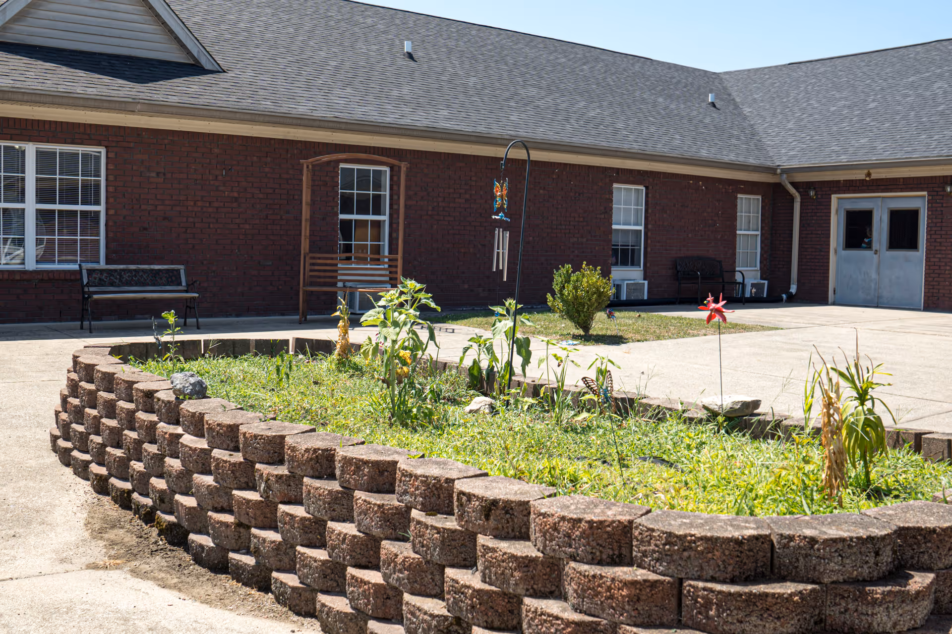 Outdoor courtyard area with a raised brick planter filled with plants and flowers. In the background, there is a brick building with several windows, benches, and a double door entrance under a clear blue sky.