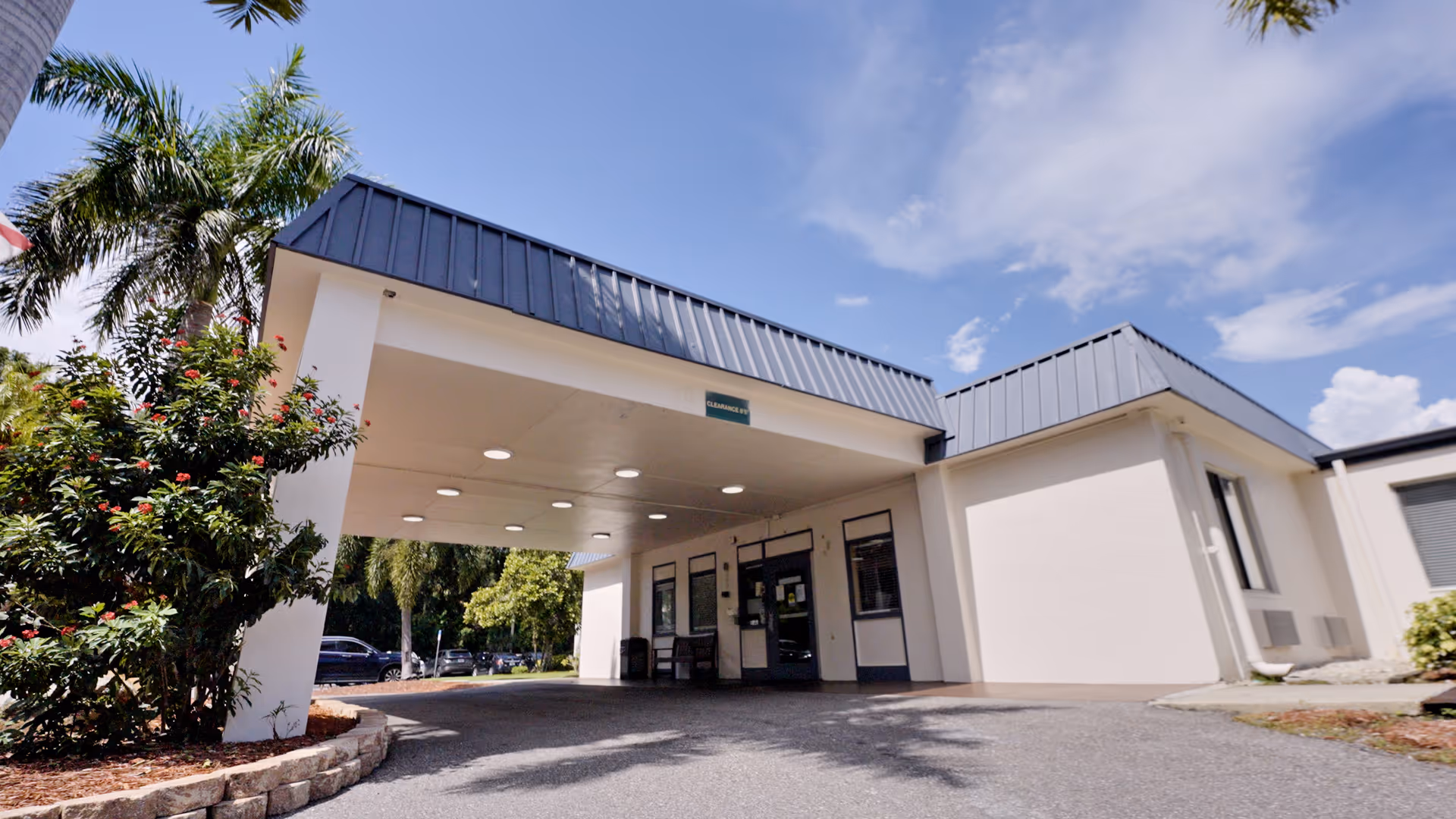 Entrance of a single-story building with a covered drop-off area, surrounded by palm trees and shrubs under a partly cloudy sky.