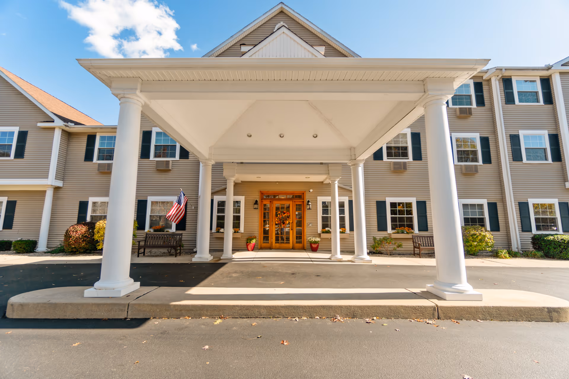 Front entrance with a covered portico supported by white columns, double wooden doors, benches, and an American flag.