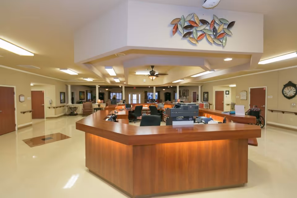 Interior view of a healthcare facility reception area with a large wooden reception desk in the center. The desk is surrounded by office chairs and office equipment. The room has beige walls, several doors, handrails along the walls, a ceiling fan, and decorative wall art above the reception desk. There is a person in a wheelchair near one of the doors on the right side.